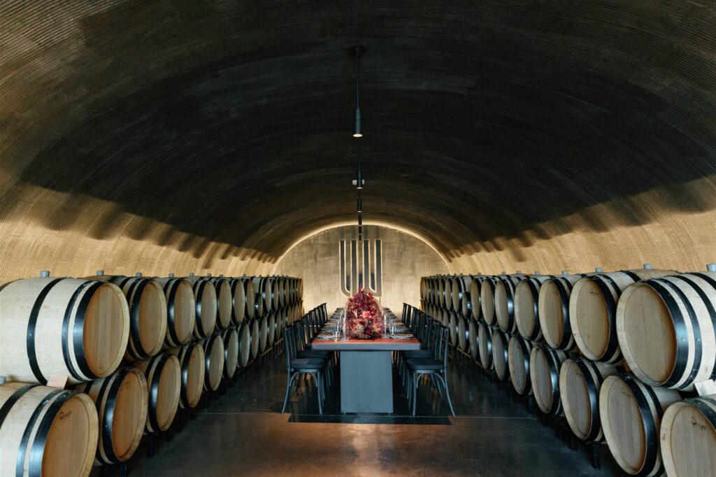 Moody rehearsal dinner tablescape with deep red florals and candlelight among wine barrels.