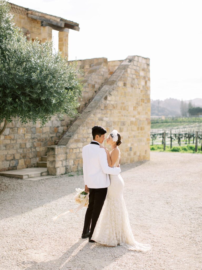 Bride and groom embracing beside a stone estate wall overlooking the vineyards, from a wine country destination wedding.
